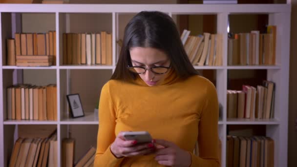 Ritratto di bella insegnante bruna in occhiali che lavora con smartphone si rivolge alla fotocamera e sorride alla biblioteca . — Video Stock