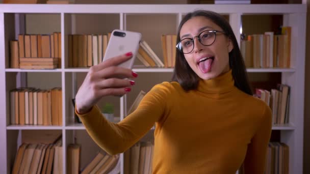 Beautiful brunette female teacher in glasses making funny selfies on smartphone showing tongue joyfully at the library. — Stock Video