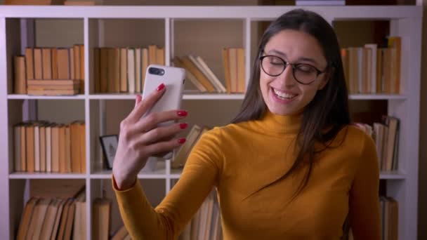 Hermosa profesora morena en gafas haciendo selfies divertidos y raros en el smartphone de la biblioteca . — Vídeos de Stock