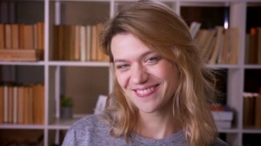 Orta yaşlı sarışın öğretmen kameraya döner ve kütüphanede son derece mutlu olmak kamera içine gülümsüyor.