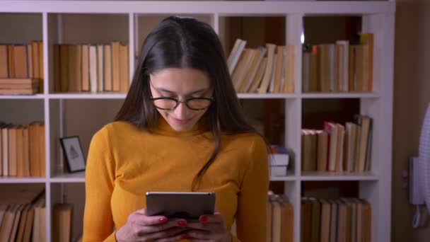 Portrait de belle enseignante brune en lunettes travaillant avec une tablette se tourne vers la caméra et sourit à la bibliothèque . — Video