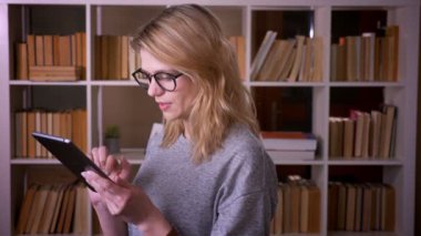 Kütüphanede yoğunlaşan tablete özenle izleyen orta yaşlı sarışın öğretmenin profil portresi.