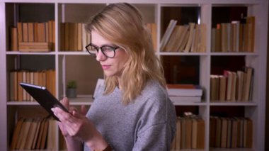 Tablet konsantre olma ile çalışan orta yaşlı sarışın öğretmenin profil portresi kütüphanede kameraya döner.