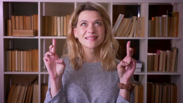 Cute middle-aged blonde teacher prays with crossed fingers to show hope into camera at the library. — Stock Video