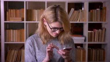 Akıllı telefon la çalışan orta yaşlı sarışın öğretmen neşeli kameraya döner ve kütüphanede gülümsüyor.