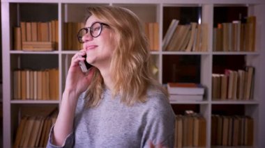 Güzel orta yaşlı sarışın öğretmen kütüphanede neşeli ve memnun olmak akıllı telefon bir voicecall sahip.