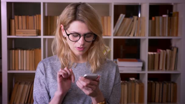 Bonita profesora rubia de mediana edad observando atentamente en el teléfono inteligente concentrado en la biblioteca . — Vídeos de Stock