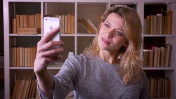 Beautiful middle-aged blonde teacher making attractive selfies using smartphone at the library. — Stock Video