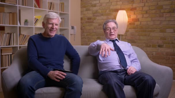 Portret van senior mannelijke vrienden zitten samen op de sofa kijken tv en discussiëren actief en vreze. — Stockvideo