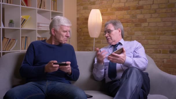 Retrato de amigos varones mayores sentados cada uno en su propio teléfono inteligente y discutiendo atenta y seriamente . — Vídeo de stock