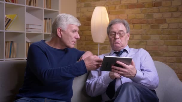 Портрет старших друзів-чоловіків, які сидять разом на дивані, витираючи фотографії на планшеті та емоційно реагуючи . — стокове відео