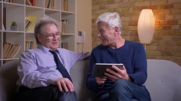 Retrato de amigos varones mayores sentados juntos en el sofá deslizando fotos en la tableta y riendo felizmente . — Vídeo de stock