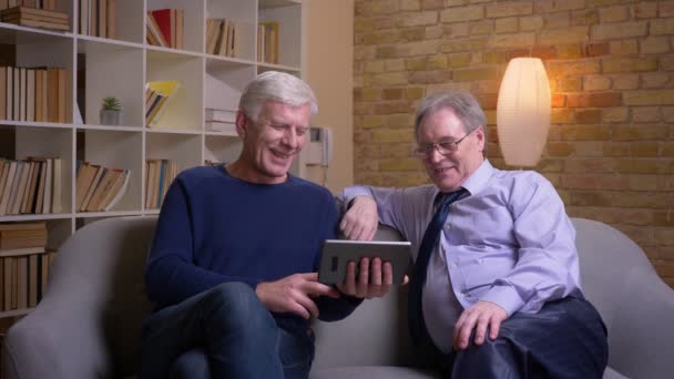 Retrato de amigos varones mayores sentados juntos en el sofá hablando en videochat en la tableta siendo feliz . — Vídeo de stock