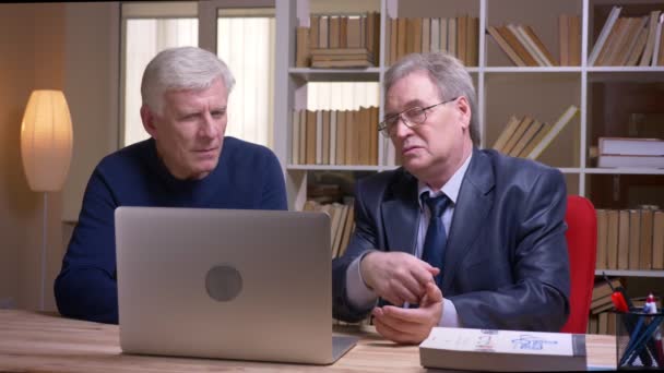 Портрет старших бізнесменів, які сидять разом за столом, працюють з ноутбуком і серйозно обговорюють проект . — стокове відео