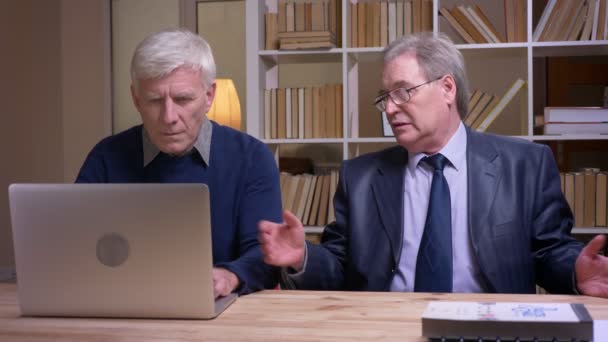 Портрет старих бізнесменів, які сидять разом за столом, працюють з ноутбуком і активно обговорюють контракт . — стокове відео