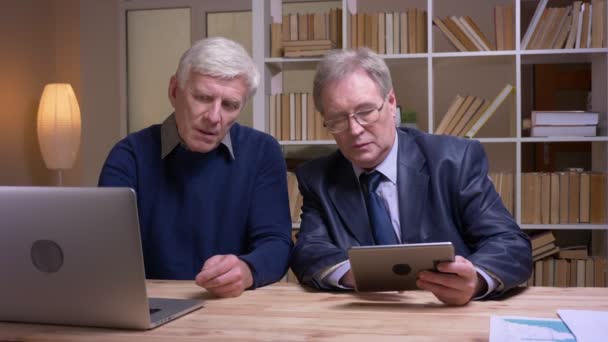 Retrato de viejos empresarios que trabajan junto con el ordenador portátil y la tableta discutiendo seriamente el proyecto . — Vídeos de Stock