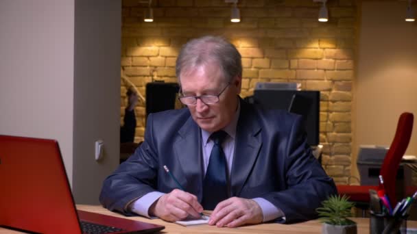 Retrato del hombre mayor en traje formal que trabaja con el ordenador portátil y escribir notas en el cuaderno en la oficina . — Vídeos de Stock