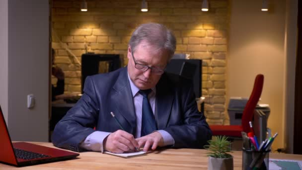 Retrato de hombre de negocios senior en traje oficial escribiendo notas en cuaderno se convierte en cámara y sonríe en la oficina . — Vídeos de Stock