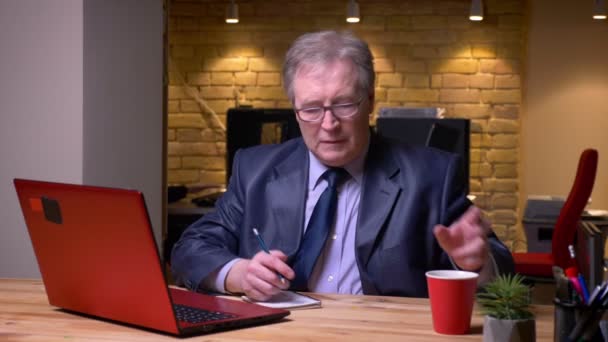 Retrato del hombre mayor en traje formal escribiendo en el portátil y escribiendo notas en el cuaderno que se concentra en la oficina . — Vídeo de stock