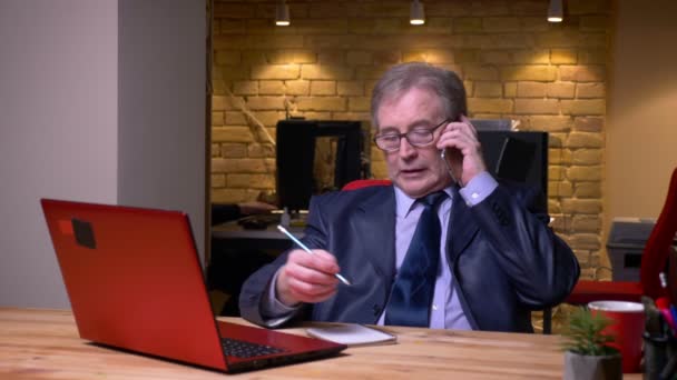 Senior-Geschäftsmann in festlichem Kostüm vor Laptop und Notizen in Notizbuch reden auf Handy im Büro. — Stockvideo