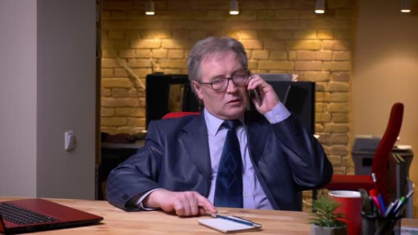 Senior affärsman i formell kostym framför laptop pratar uppmärksamt på mobiltelefon i Office. — Stockvideo