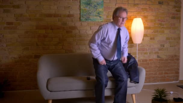 Portrait of senior businessman in costume sits down on sofa and switches on the TV after hard working day. — Stock Video