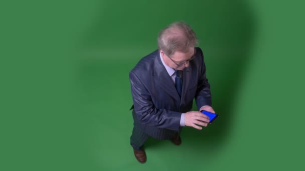 Full-Height top shot van senior man draait op app en toont blauw scherm van de telefoon op groene Chromakey achtergrond. — Stockvideo