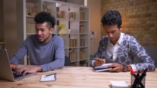 Närbild shoot av afroamerikanska och indiska manliga studenter lärande tillsammans. Man skriver på den bärbara datorn medan en annan tar anteckningar — Stockvideo