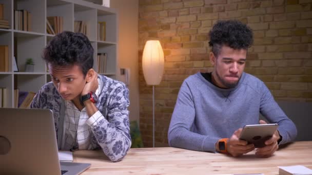 Sesión de primer plano del estudiante indio que trabaja duro aprendiendo usando la computadora portátil y la mensajería afro-americana felizmente en la tableta que está sentada al lado en la biblioteca — Vídeos de Stock