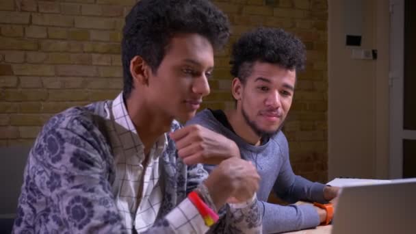 Gros plan sur des étudiants indiens et afro-américains utilisant les ordinateurs portables et discutant d'un projet heureux de sourire assis à l'intérieur de la bibliothèque ensemble — Video