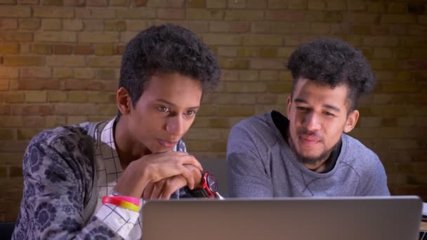 Syuting closeup muda India dan african american teman laki-laki muda menonton film di laptop dengan kegembiraan duduk di dalam ruangan — Stok Video