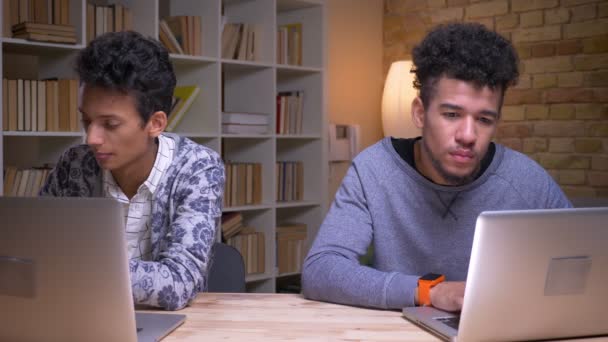 Sesión de primer plano de estudiantes varones indios y afroamericanos usando las computadoras portátiles y aprendiendo en línea sentados en el interior de la biblioteca juntos — Vídeos de Stock