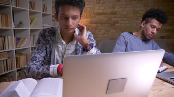 Syuting closeup dengan kamera bergerak dari India untuk Afrika Amerika laki-laki siswa menggunakan laptop dan belajar online duduk di dalam ruangan di perpustakaan — Stok Video