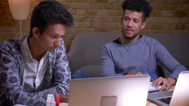 Tiro de perto de estudantes indianos e afro-americanos do sexo masculino usando os laptops e discutindo um projeto universitário sentado dentro da biblioteca juntos — Vídeo de Stock