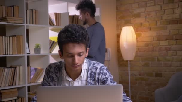 Närbild shoot av två kulturellt olika studenter lärande i kollegiet biblioteket inomhus. Indiska manliga studerar på nätet på den bärbara datorn medan afrikansk amerikansk man rusing tabletten på bakgrunden — Stockvideo