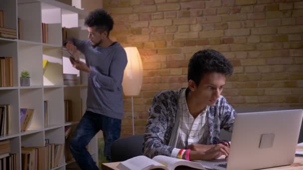 Съемки крупным планом двух культурно разнообразных учащихся, обучающихся в школьной библиотеке в помещении. Индийский мужчина учится онлайн на ноутбуке, в то время как африканский американец использует планшет на заднем плане — стоковое видео