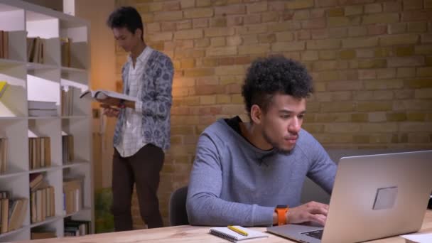 Foto ravvicinata di due studenti etnicamente diversi che studiano insieme in biblioteca. Uomo afroamericano che utilizza il computer portatile mentre l'uomo indiano legge il libro sullo sfondo — Video Stock