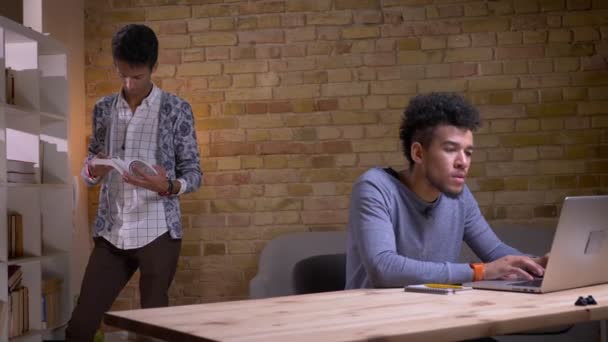 Closeup shoot av två etniskt olika studenter som studerar tillsammans i Universitetsbiblioteket. Afroamerikansk hane som använder den bärbara datorn medan indisk man läser boken på bakgrunden — Stockvideo