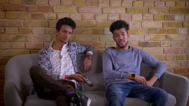 Syuting jarak dekat dari dua teman laki-laki muda menonton TV bersama-sama dengan kegembiraan duduk di sofa di dalam ruangan di sebuah apartemen nyaman — Stok Video
