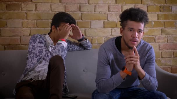 Närbild shoot av två unga manliga vänner tittar på en skräckfilm på TV tillsammans få ett hopp skrämma sitter på soffan inomhus i en mysig lägenhet — Stockvideo