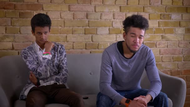 Syuting jarak dekat dari dua teman laki-laki muda menonton drama sedih di TV bersama-sama duduk di sofa di dalam ruangan di sebuah apartemen nyaman — Stok Video