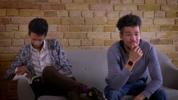 Съемки крупным планом двух молодых друзей-мужчин, сидящих вместе на диване в уютной квартире. Индийский мужчина учится и отвлекается на счастливого африканского американца, смотрящего телевизор — стоковое видео