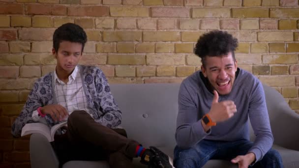 Střílejí dva mladí mužští přátelé sedícího na gauči společně v útulném bytě. Indický muž se učí a rozptylována se smějící afroameričanovi, který se dívá na televizi — Stock video