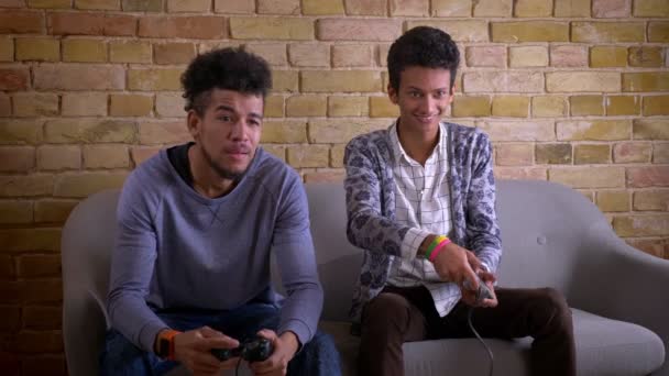 Zbliżenie strzelać African American i Indian męskich przyjaciół grając w gry wideo razem siedzi na kanapie w pomieszczeniu. Jeden wygrywa nad innym triumfem jest szczęśliwy — Wideo stockowe