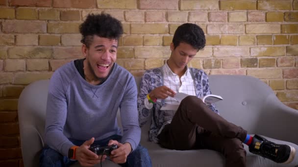 Close-up shoot van twee jonge mannelijke vrienden zittend op de Bank samen binnen in een gezellig appartement. Indian man studeert terwijl Happy African American man speelt video games — Stockvideo