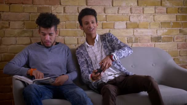 Съемки крупным планом двух молодых друзей-мужчин, сидящих на диване вместе в помещении в уютной квартире.Африканский американец изучает и отвлекается на счастливый индийский мужчина играет в видеоигры — стоковое видео