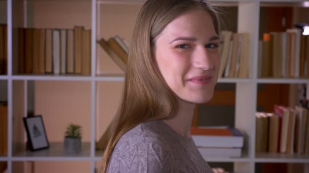 Крупный план портрета молодой привлекательной студентки, поворачивающейся и счастливо смотрящей на камеру в библиотеке колледжа — стоковое видео