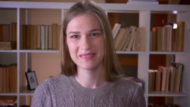 カメラを見て、屋内の大学図書館で楽しそうに微笑む若い魅力的な女子学生のクローズアップポートレート — ストック動画