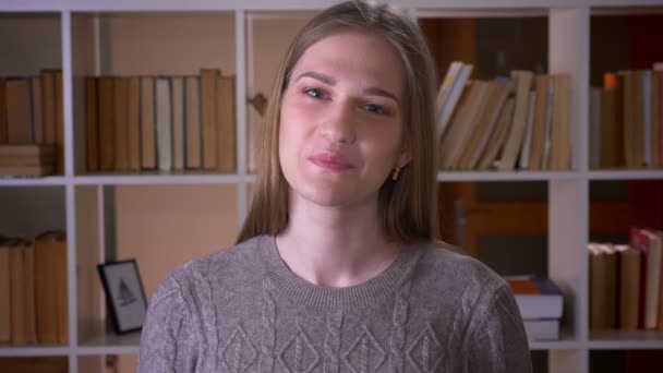 Närbild porträtt av unga attraktiva kvinnlig student tittar på kamera och leende lyckligt med spänning i kollegiet biblioteket inomhus — Stockvideo