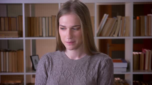 Retrato de cerca de una joven estudiante atractiva mirando a la cámara sonriendo con timidez en la biblioteca de la universidad en el interior — Vídeos de Stock